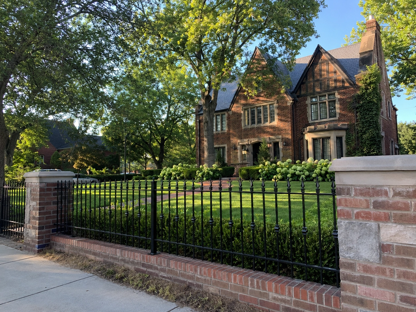 A spear-top ornamental iron fence with red-brick columns running along the front of an upscale brick Tudor home framed by mature trees, built by a fence company near me in Midtown Tulsa, OK.